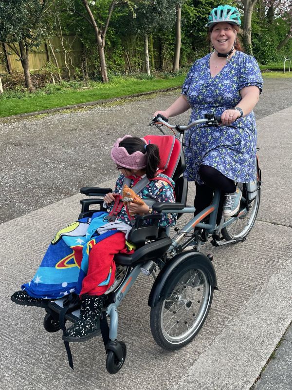 Kate on a bicycle, pushing her daughter, Mira's wheelchair in front of her.